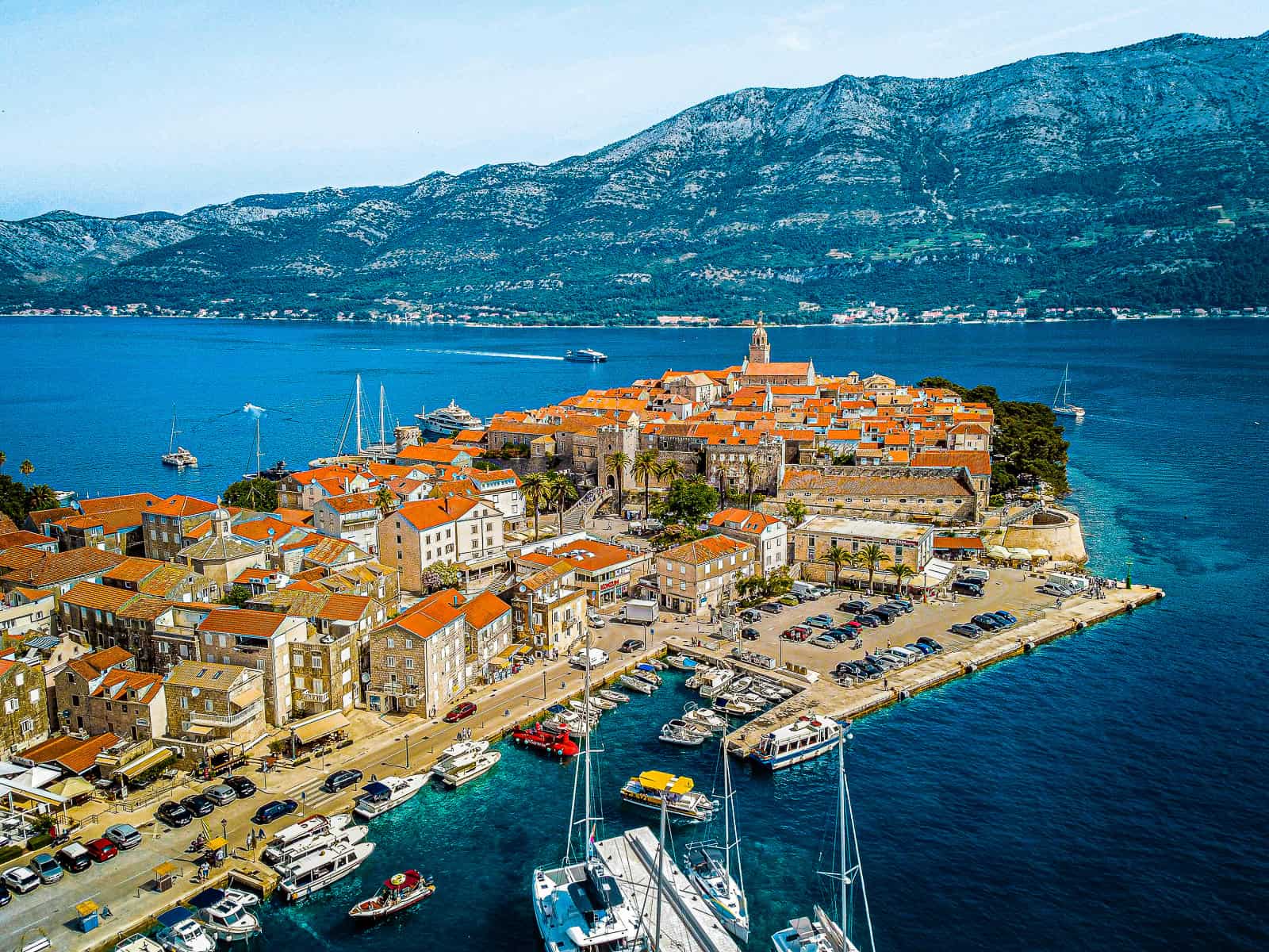 An aerial photo of old town Korcula, a popular day trip from Dubrovnik. It shows the old town surrounded by water on three sides with mountains in the background.