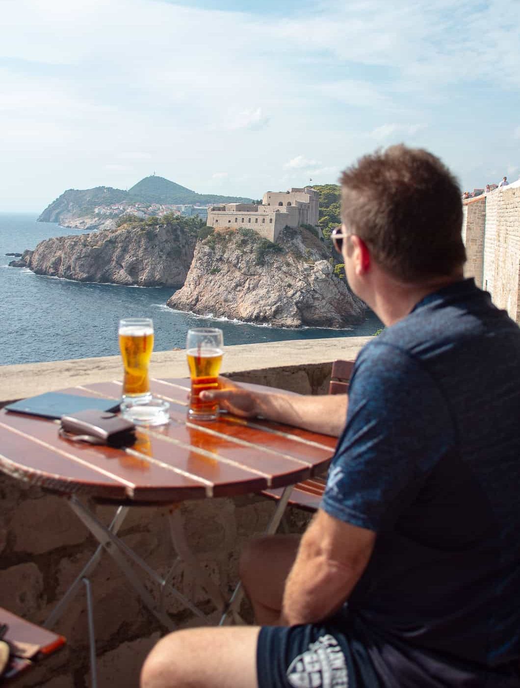A photo of a man sitting at a table on top of the Dubrovnik city walls with a beer while looking away from the camera at a view of Fort Lovrijenac in the distance.
