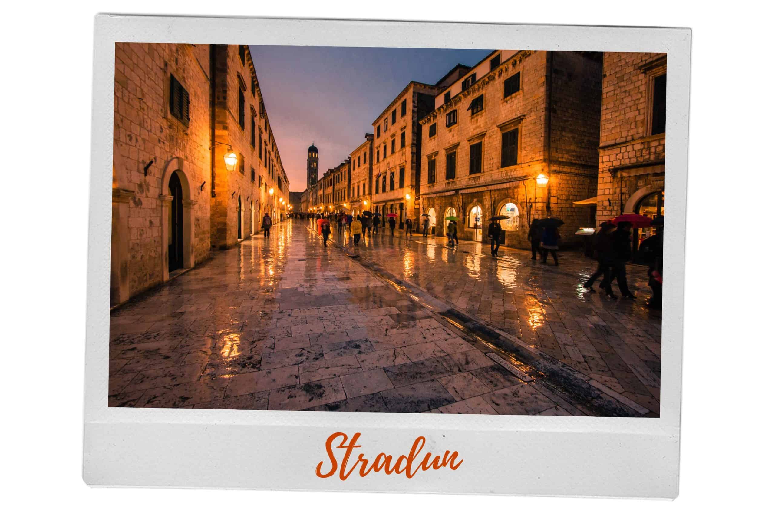 A picture of Stradun, the main street in Dubrovnik's Old Town, in the rain at night. The buildings on both sides of street are illuminated by the lights on the buildings. It shows people making their way down the street with umbrellas.