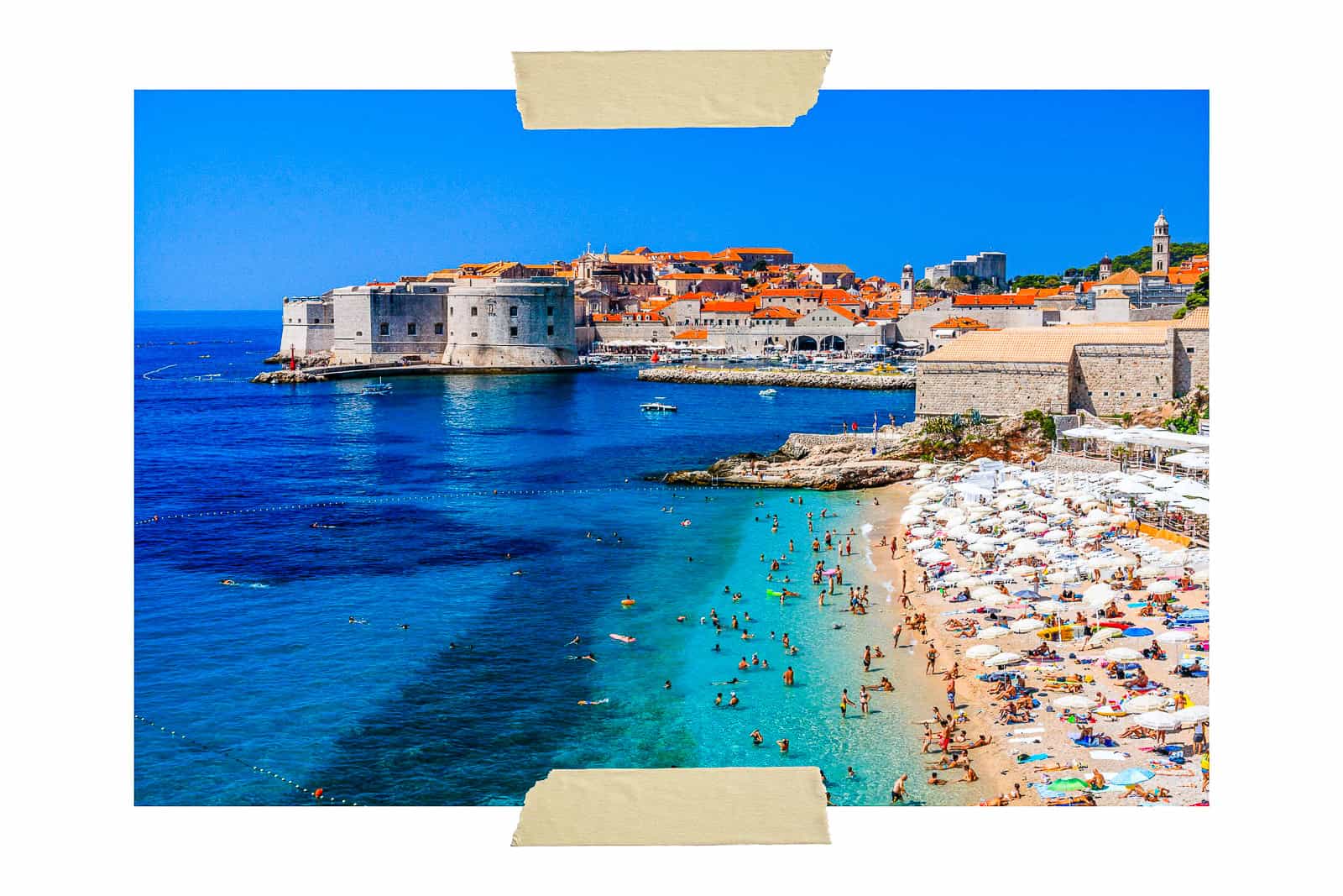 A picture of Banje Beach in Dubrovnik with the Old Town in the background and the turquoise Adriatic Sea to the left. The beach is to right with people laying in the sun and some gathering in the water. Banje is located in Ploce neighborhood which is where to stay in Dubrovnik if you're a couple looking for a luxury stay.