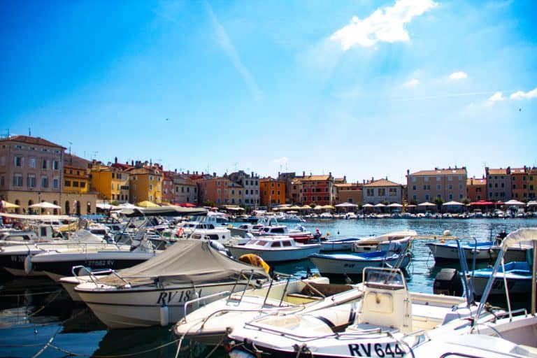 A beautiful picture of Rovinj, Croatia in early September with boats anchored in the harbor. In the background there are colorful buildings and restaurant umbrellas lining the harbor.