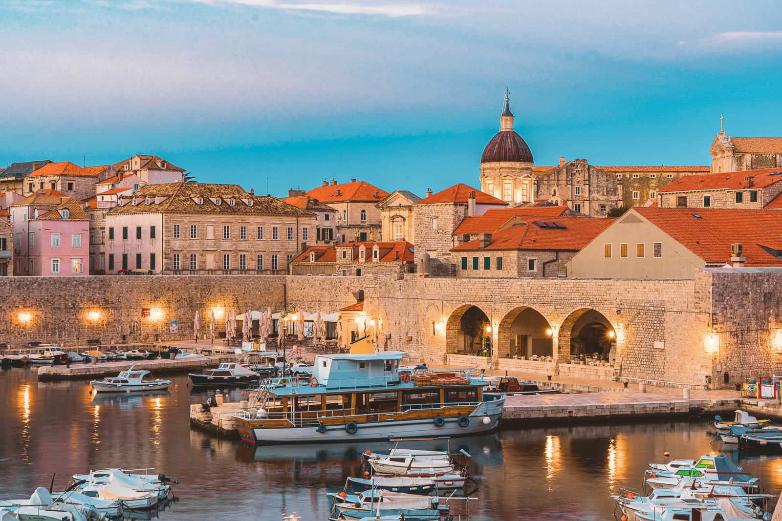 A picture of Dubrovnik's Old Town harbor showing several boats anchored in the water. The view shows a section of the city walls and the red roofed buildings behind it. The walls are illuminated by lanterns that line the walkway around the harbor.