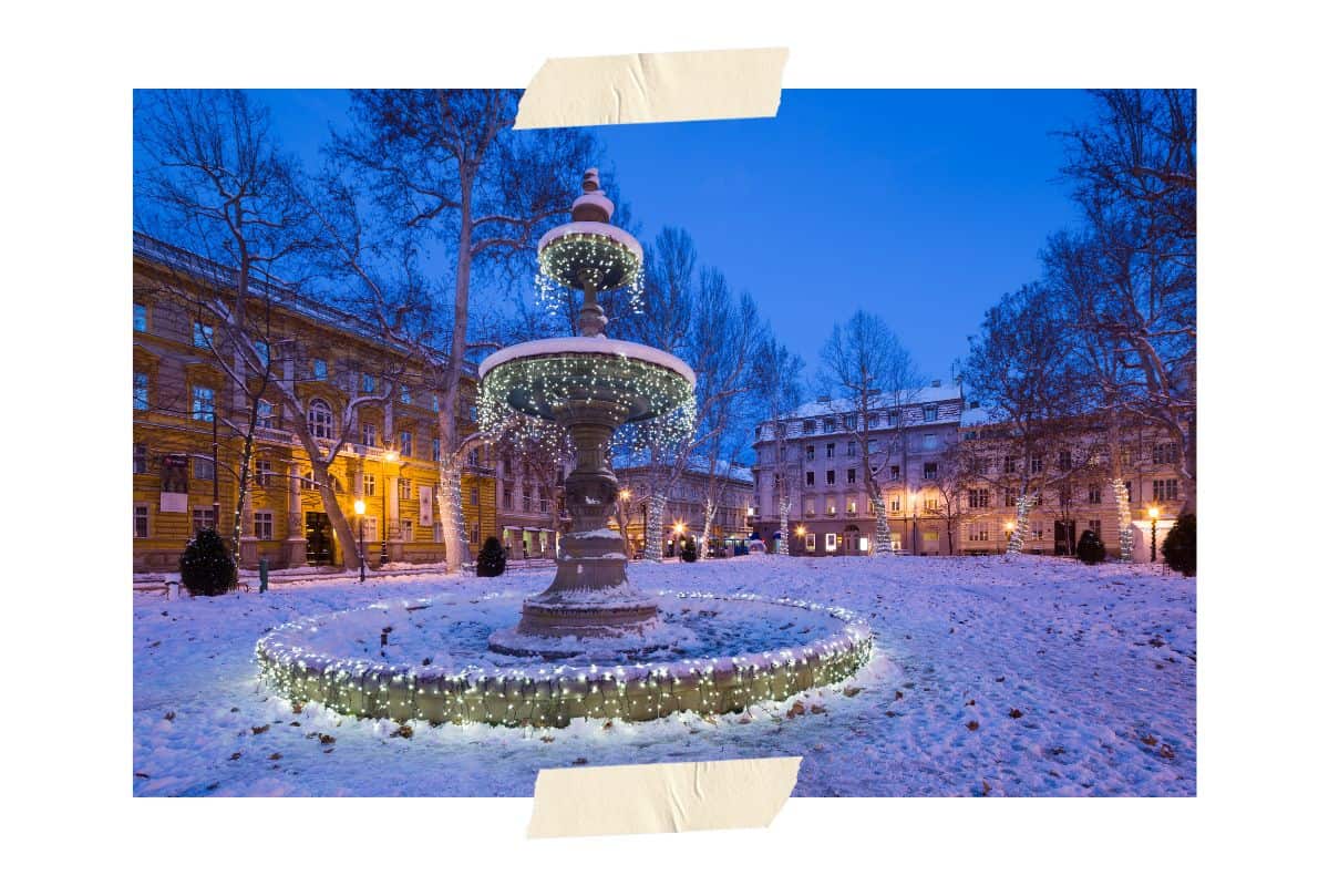 Nighttime in Zagreb during winter. A fountain decorated with Christmas lights and the ground dusted with snow in front of buildings with snow covered roofs.