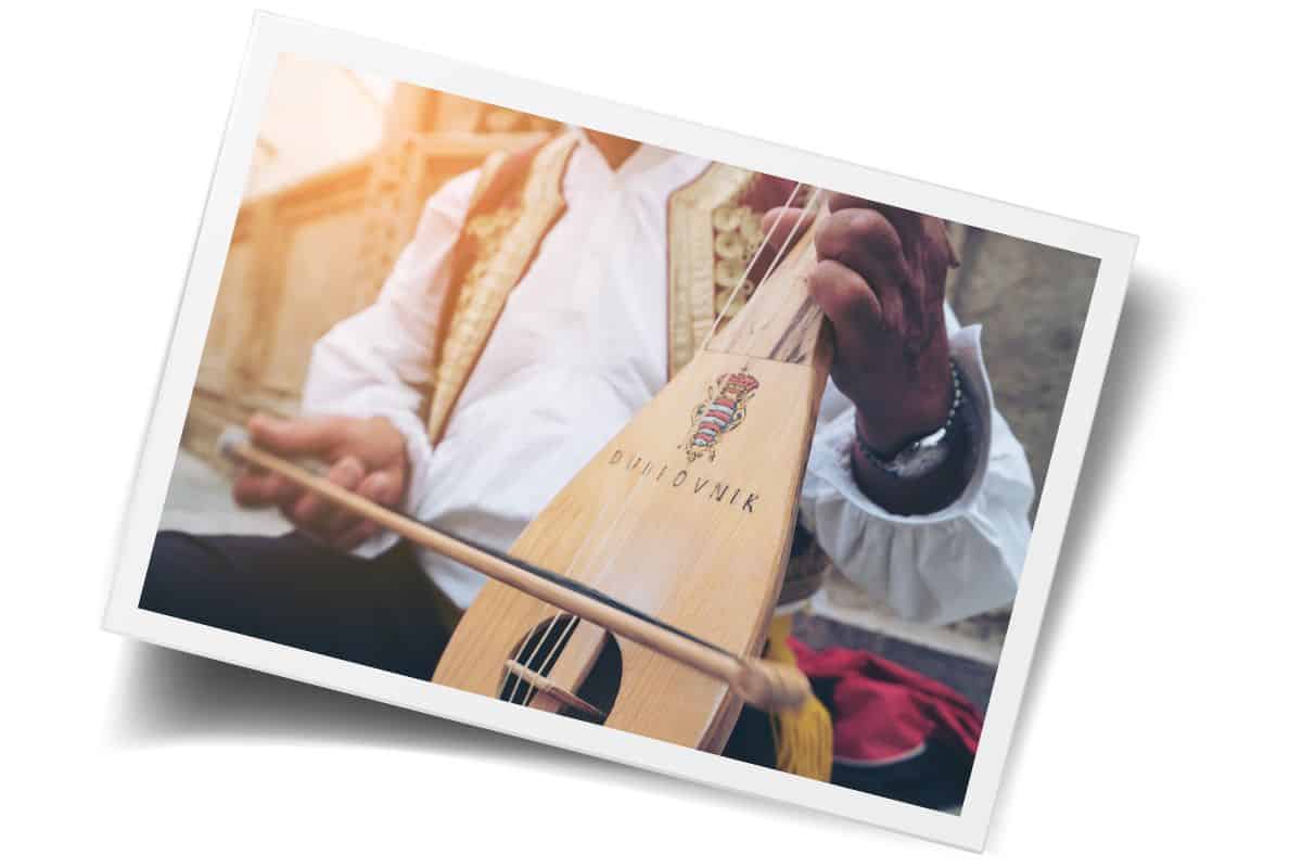 Traditional Croatian street musician playing a lijerica in Dubrovnik's Old Town, wearing folk costume, with the word "Dubrovnik" engraved on the instrument.
