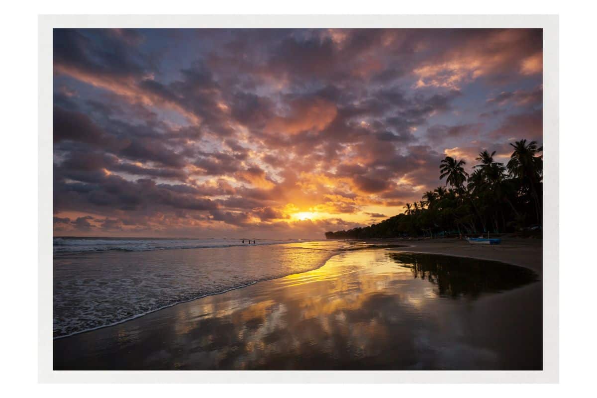 Dramatic sunset over a Costa Rica beach with golden light breaking through dark clouds. The sun's reflection creates a golden path across wet sand, with palm trees silhouetted along the shoreline.