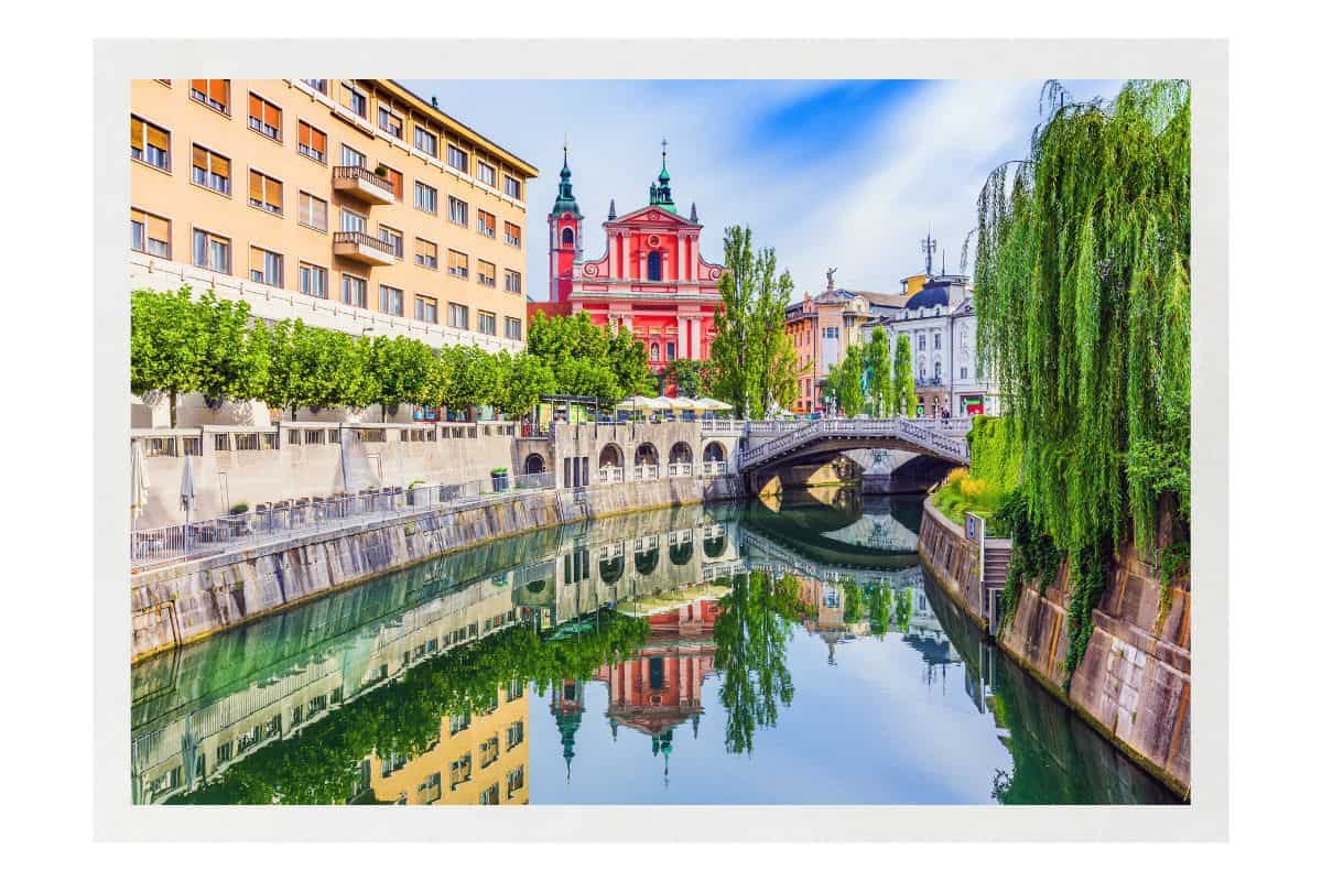 Ljubljana river canal with colorful buildings, pink church, stone bridge, and green reflections in Slovenia's capital city.