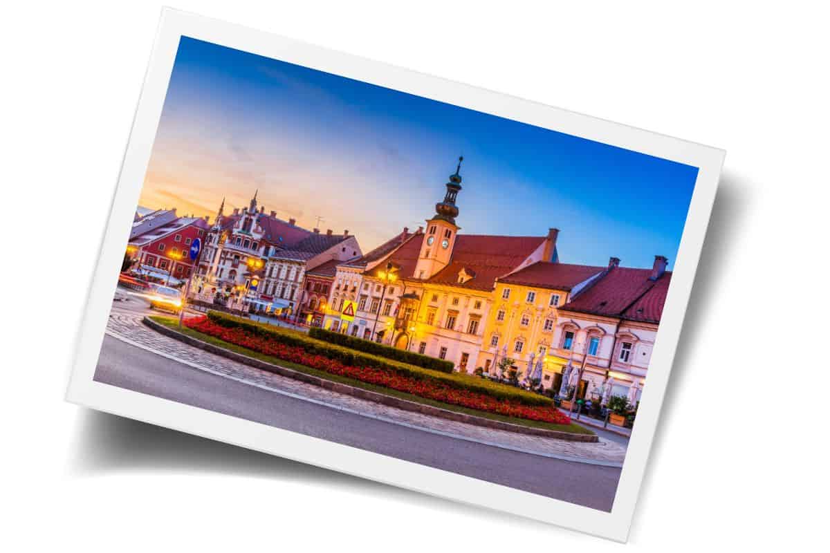 Historic town square in Slovenia at sunset with illuminated yellow and red buildings, clock tower, and flower gardens during the best visiting season.