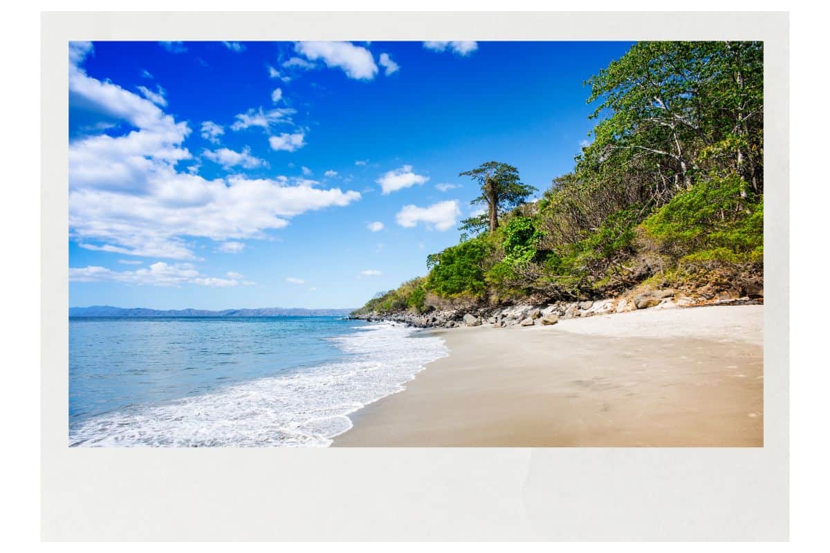 Secluded beach with gentle waves, white sand, and rocky coastline backed by green forest under blue skies in Costa Rica.