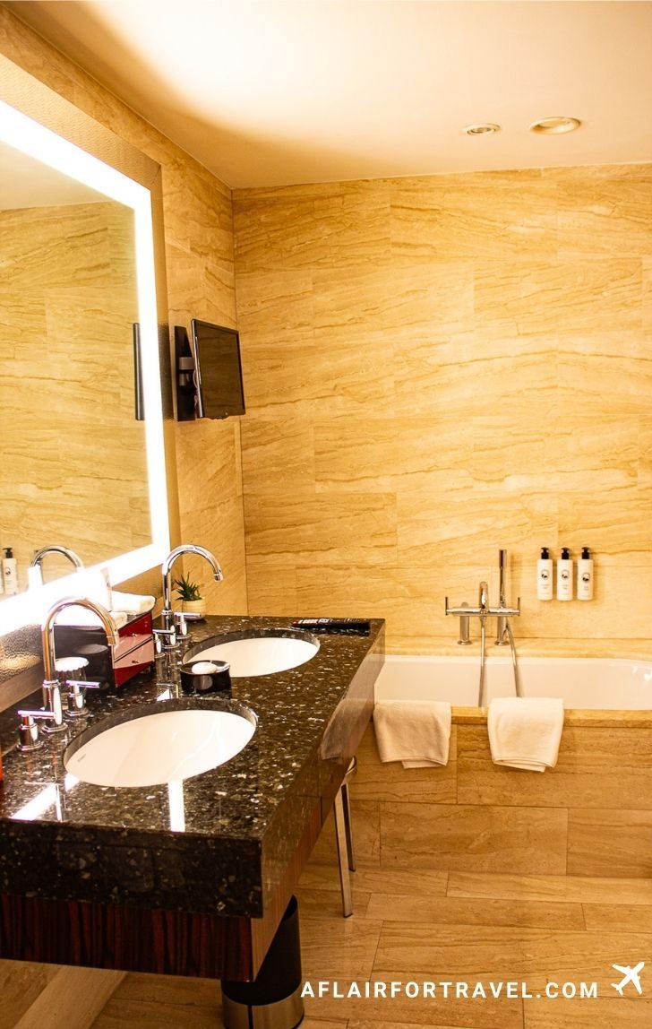 Luxurious bathroom with beige marble walls, black granite double vanity, soaking tub, and backlit mirror at Mandarin Oriental Prague