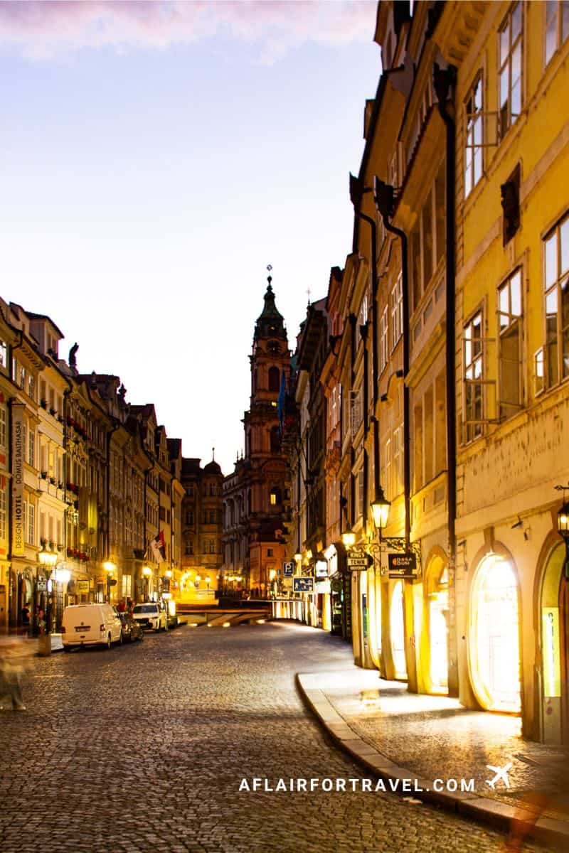 Illuminated cobblestone street in the Mala Strana neighborhood in Prague at dusk with historic buildings and church tower in the background