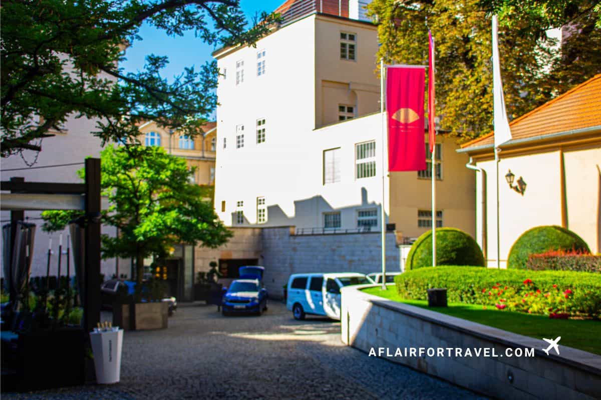 Entrance courtyard of Mandarin Oriental Prague with manicured gardens, hotel flag, and cobblestone driveway