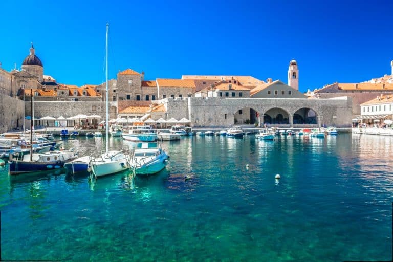 Small boats and sailboats docked in Dubrovnik’s Old Port, with clear turquoise water in the foreground and the stone city walls and terracotta rooftops of the Old Town under a bright blue sky.