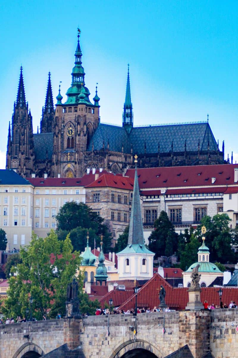 Prague Castle and St. Vitus Cathedral towers rising above red-tiled rooftops with Charles Bridge in foreground at dusk
