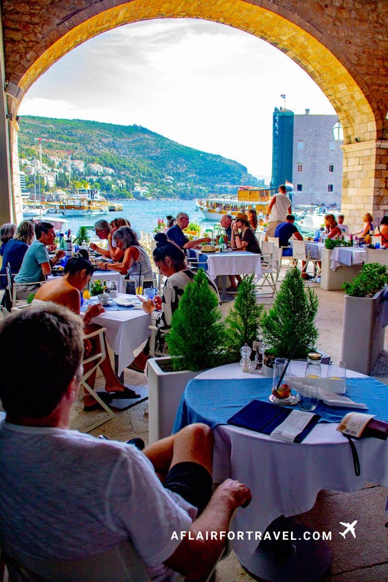 Diners at Gradska kavana Arsenal Restaurant seated under a stone archway with harbor views of boats, hillside, and Dubrovnik's fortifications in the background.