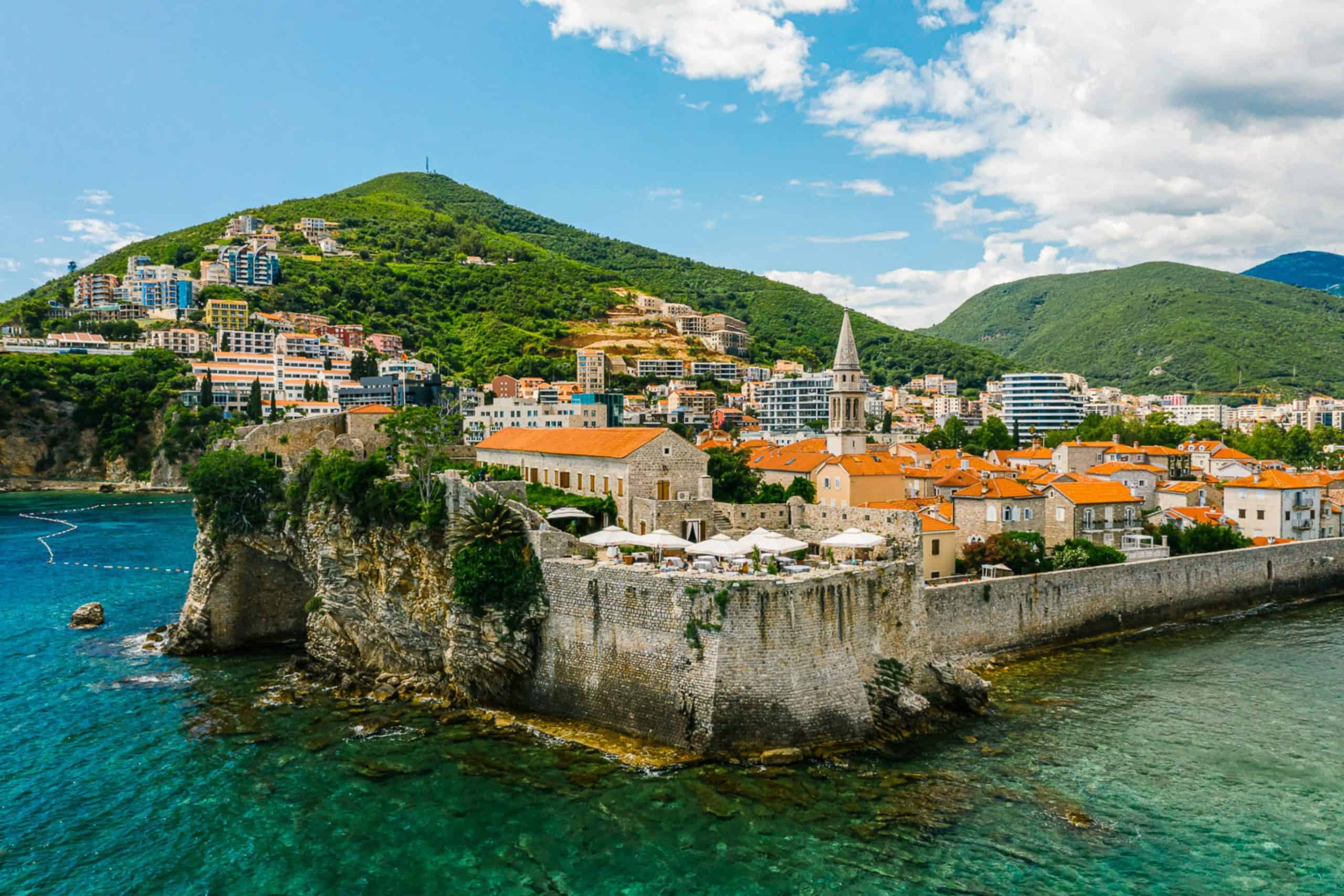 Aerial view of Budva Old Town in Montenegro, with stone city walls, orange rooftops, turquoise Adriatic Sea, and green hills in the background.