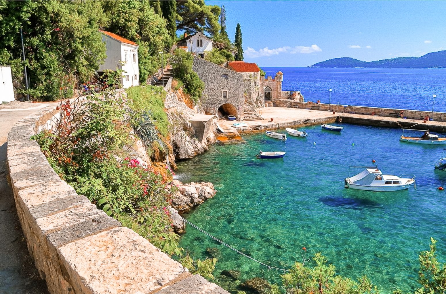 Trsteno village harbor near Dubrovnik, with stone buildings, a small marina, clear turquoise water, and boats along the Adriatic coast.