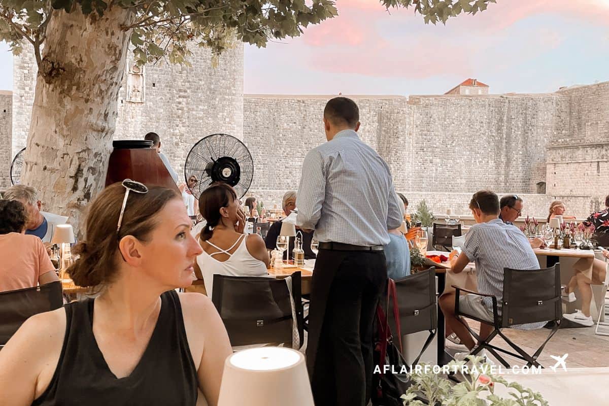 Woman sitting at a table under a tree at Dubrovka 1836 in Dubrovnik with a view of the old city walls at sunset in the background