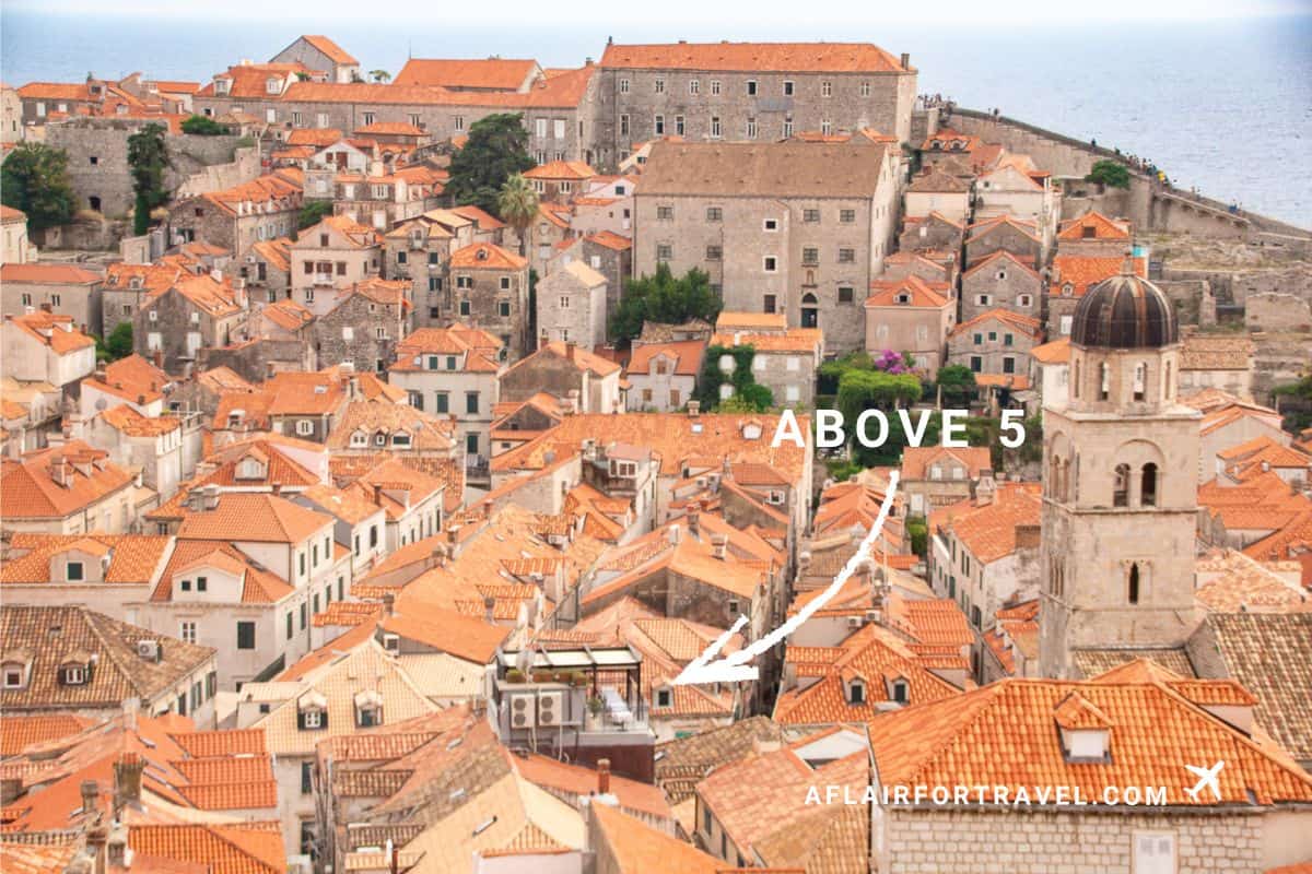 Aerial view of Dubrovnik's old town showing terracotta rooftops, stone buildings, and a church bell tower, with Above 5 restaurant marked among the rooftops near the Adriatic Sea.