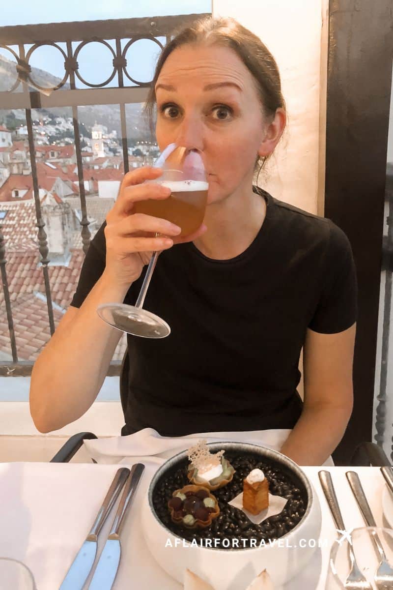 Woman sipping sangria at Above 5 Rooftop Restaurant with an elegant food plate and Dubrovnik's terracotta rooftops visible in the background at twilight.