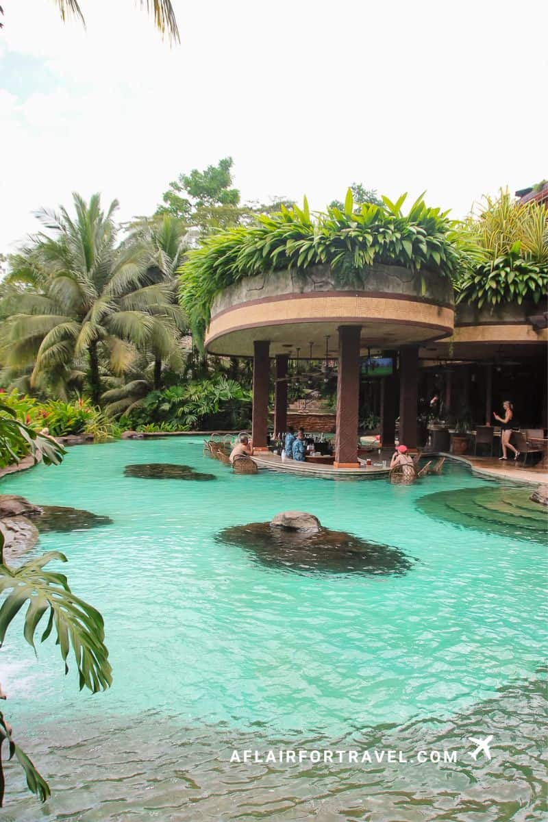 Tropical resort pool at The Springs Resort with turquoise water and swim-up bar featuring living green roof, surrounded by palm trees and lush vegetation in Costa Rica