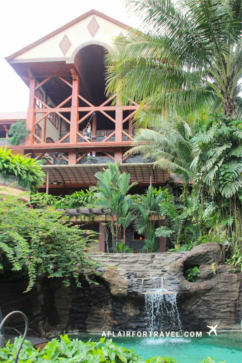 Multi-level Costa Rican resort building with wooden balconies surrounded by palm trees and tropical gardens, with natural rock waterfall cascading into turquoise pool below