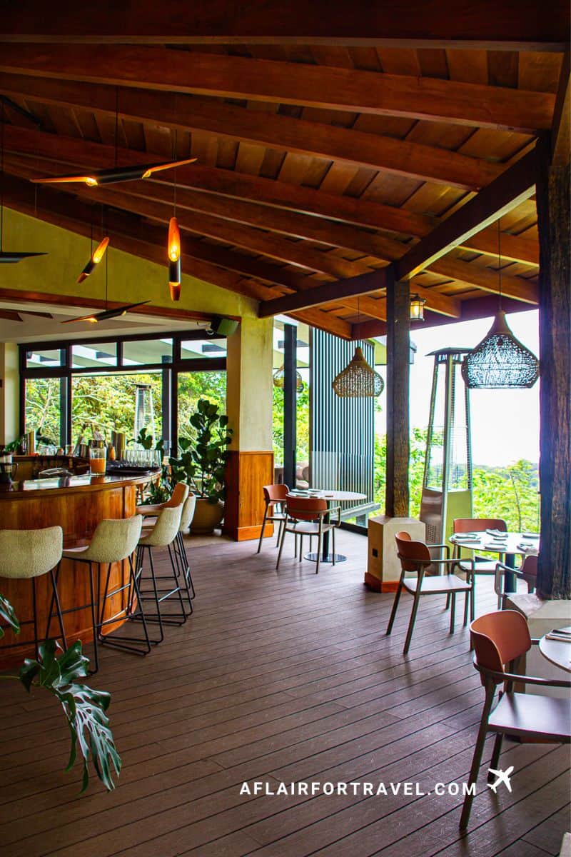 Open-air restaurant and bar at Koora Hotel with exposed beam ceiling, wooden floors, modern pendant lighting, and floor-to-ceiling windows overlooking Monteverde cloud forest