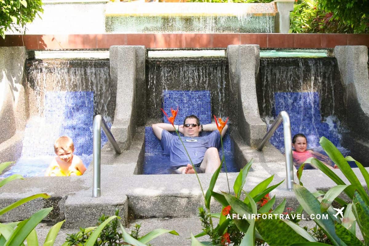 Family relaxing in three adjacent hot spring chairs with blue mosaic tile and waterfall features at a Costa Rican thermal spa resort