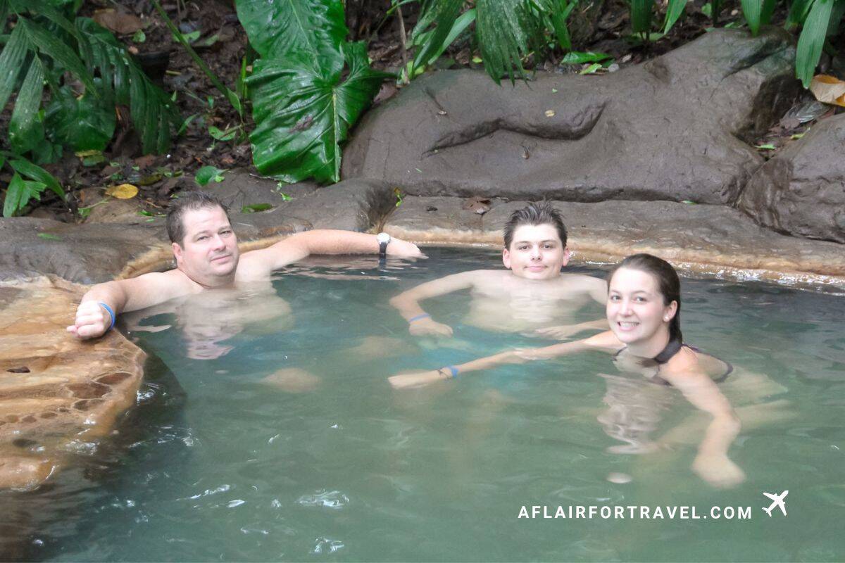 Three people relaxing in natural hot spring pool with mineral-rich turquoise water and rock edges, surrounded by tropical plants in Costa Rica