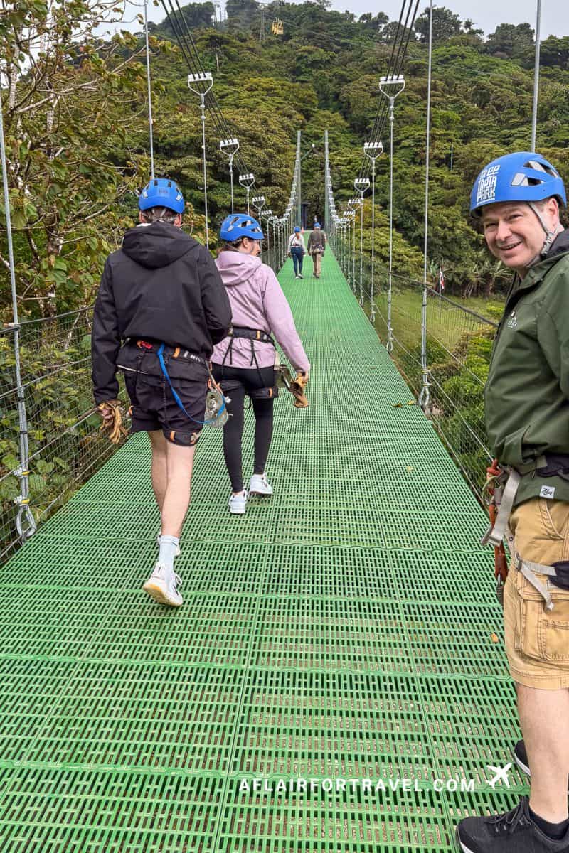 Tourists wearing blue helmets and safety harnesses walking across green suspension bridge high above cloud forest canopy in Monteverde, Costa Rica