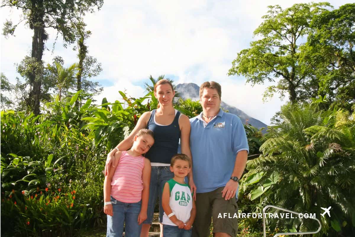 Family of four posing in front of Arenal Volcano in Costa Rica, surrounded by lush tropical vegetation including palm trees and banana plants, with the volcanic peak visible through clouds in the background