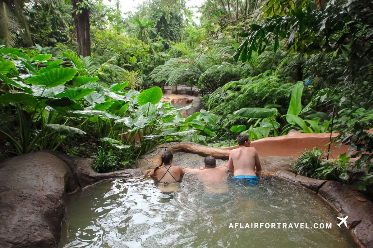 Family soaking in a rainforest hot spring surrounded by lush tropical plants in Arenal, Costa Rica.