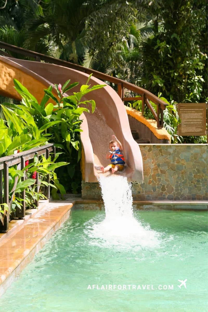 Child sliding into a warm mineral pool at Tabacón Resort hot springs surrounded by tropical greenery in Costa Rica