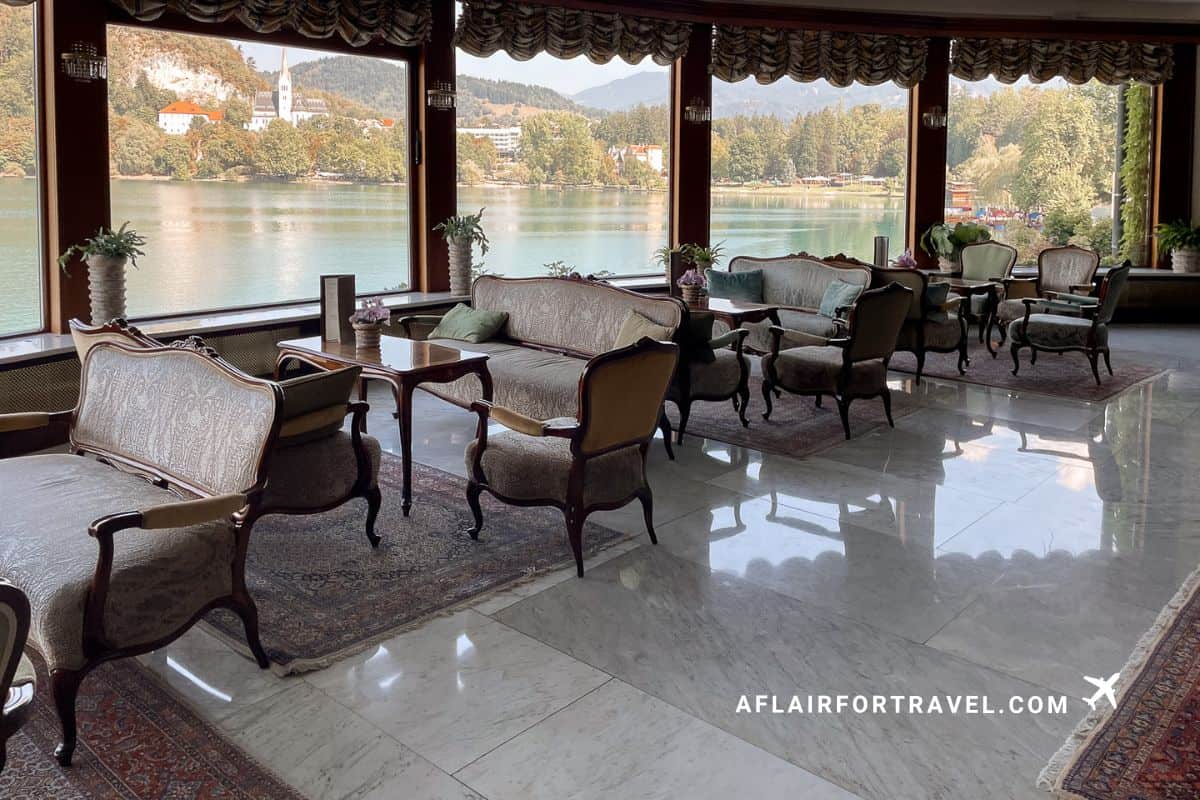 Elegant lounge at Grand Hotel Toplice featuring vintage-style furniture and floor-to-ceiling windows overlooking Lake Bled's turquoise waters and surrounding mountains
