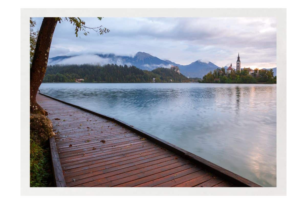 Wooden boardwalk extending along the turquoise waters of Lake Bled, with the island church and Julian Alps mountains covered in a mist visible in the distance