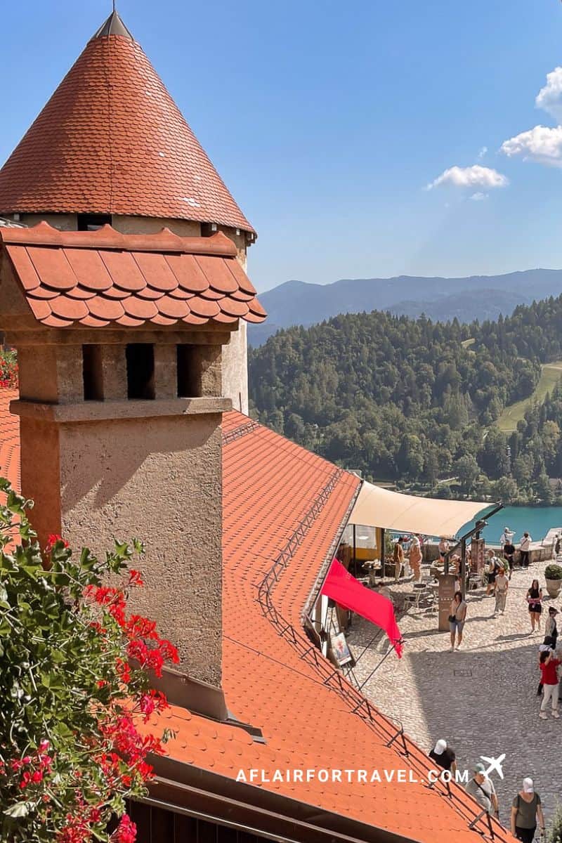 View from Bled Castle showing red-tiled tower, courtyard with visitors, and panoramic vista of the lake and Julian Alps
