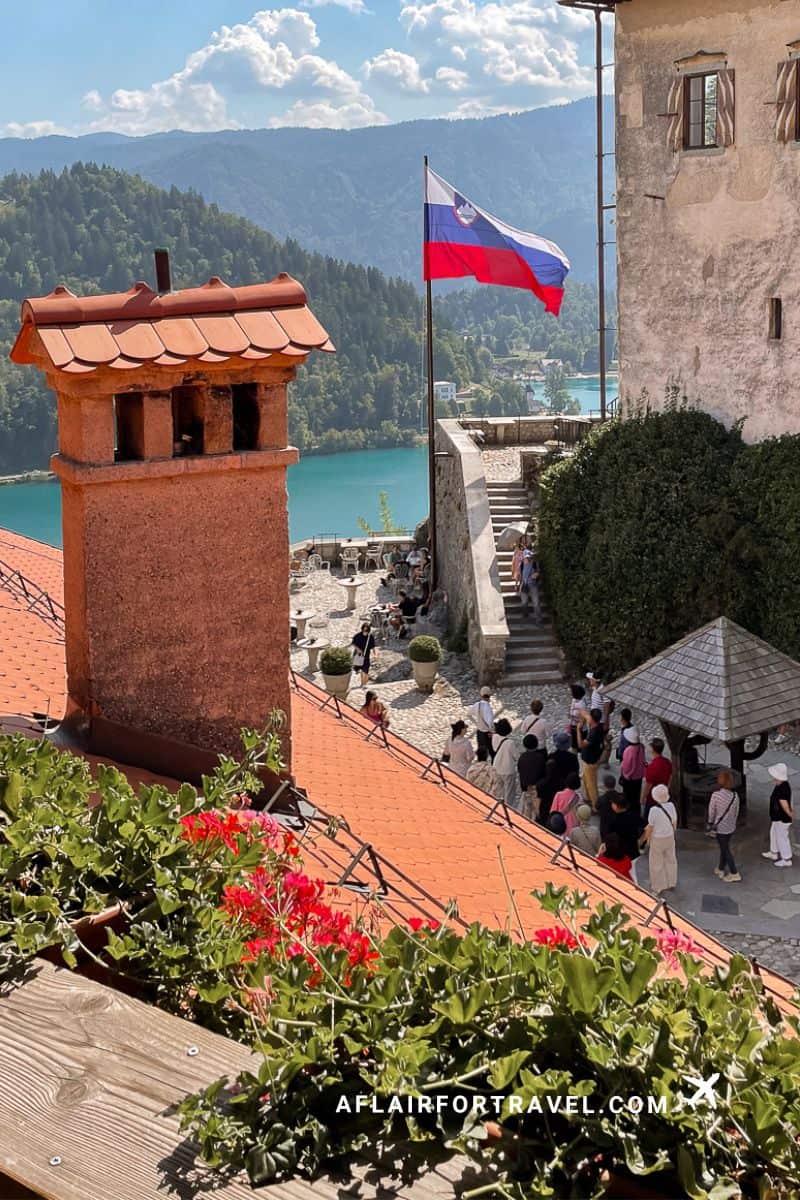 Slovenian flag flying above Bled Castle's terracotta rooftops with scenic overlook of the turquoise lake and forested mountains