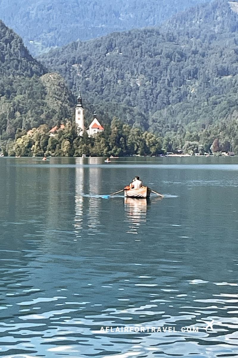 Couple rowing a wooden boat on the calm lake waters with Bled Island church and bell tower visible among the tree-covered hills