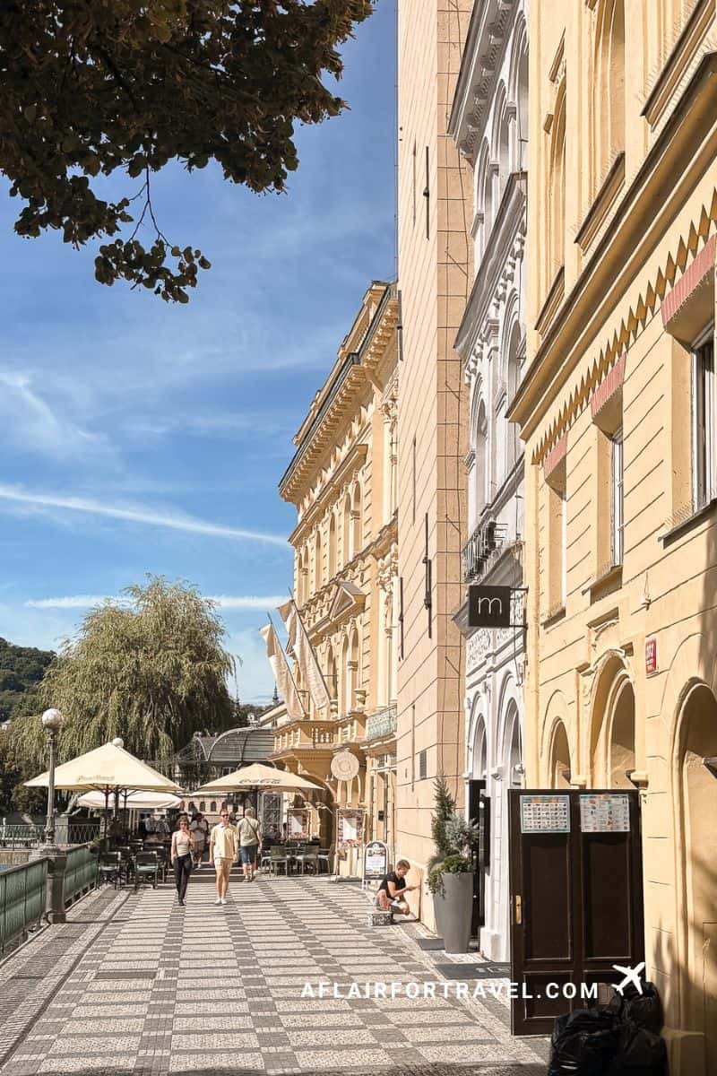 Sunny pedestrian street in Prague's Old Town with golden neoclassical buildings, outdoor cafe umbrellas, patterned pavement, and people strolling under blue sky