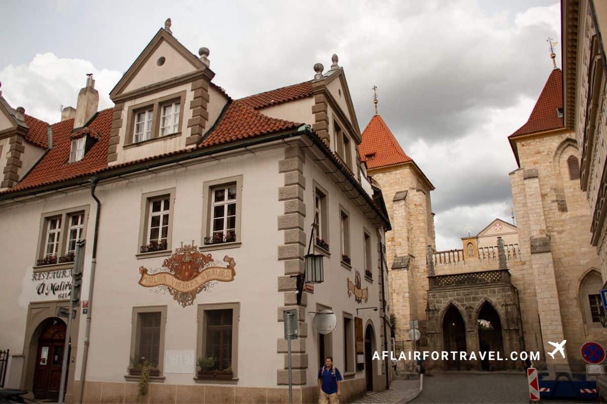 Historic Mala Strana street scene with white building featuring traditional painted house sign, terracotta tile roofs, stone tower with conical roof, and cobblestone street