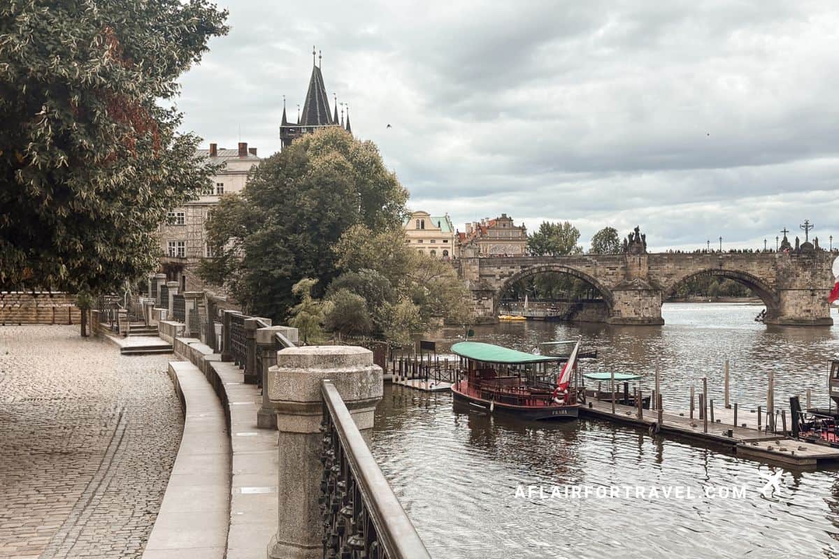 Strolling along the Vltava River waterfront in Prague is completely free with riverboats docked at pier, Charles Bridge stone arches in background, Gothic towers, and tree-lined embankment under dramatic cloudy sky