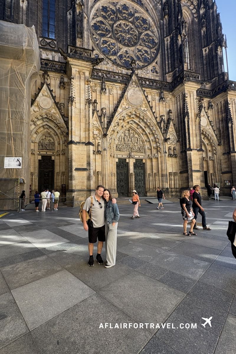 Couple standing in front of St. Vitus Cathedral's ornate Gothic entrance in Prague Castle, featuring pointed arches, rose window, and intricate stone carvings, which is free to explore the exterior