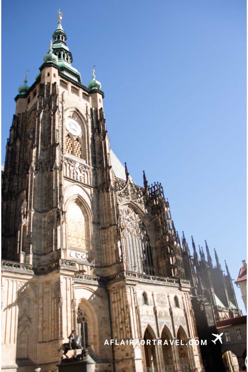 Exterior of St. Vitus Cathedral's Great South Tower with distinctive green copper domes, Gothic architecture, clock face, and flying buttresses against blue sky in Prague which is free to admire from the outside
