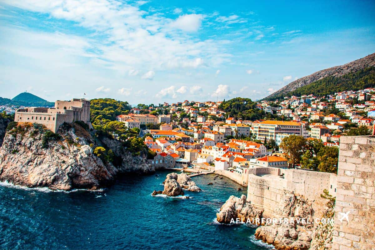 Panoramic view of Dubrovnik's Old Town with orange terracotta roofs, medieval stone walls and fortifications meeting the deep blue Adriatic Sea, with Lovrijenac Fortress on the cliff and hillside in the background, one of the main reasons it's worth visiting Croatia
