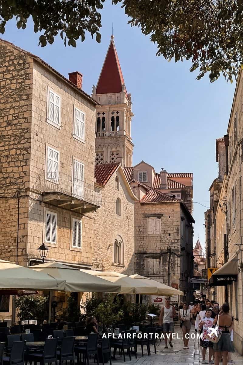 Historic stone street in Trogir's UNESCO old town, Croatia, with limestone buildings, outdoor café umbrellas, and distinctive red-roofed church bell tower rising above the Dalmatian architecture