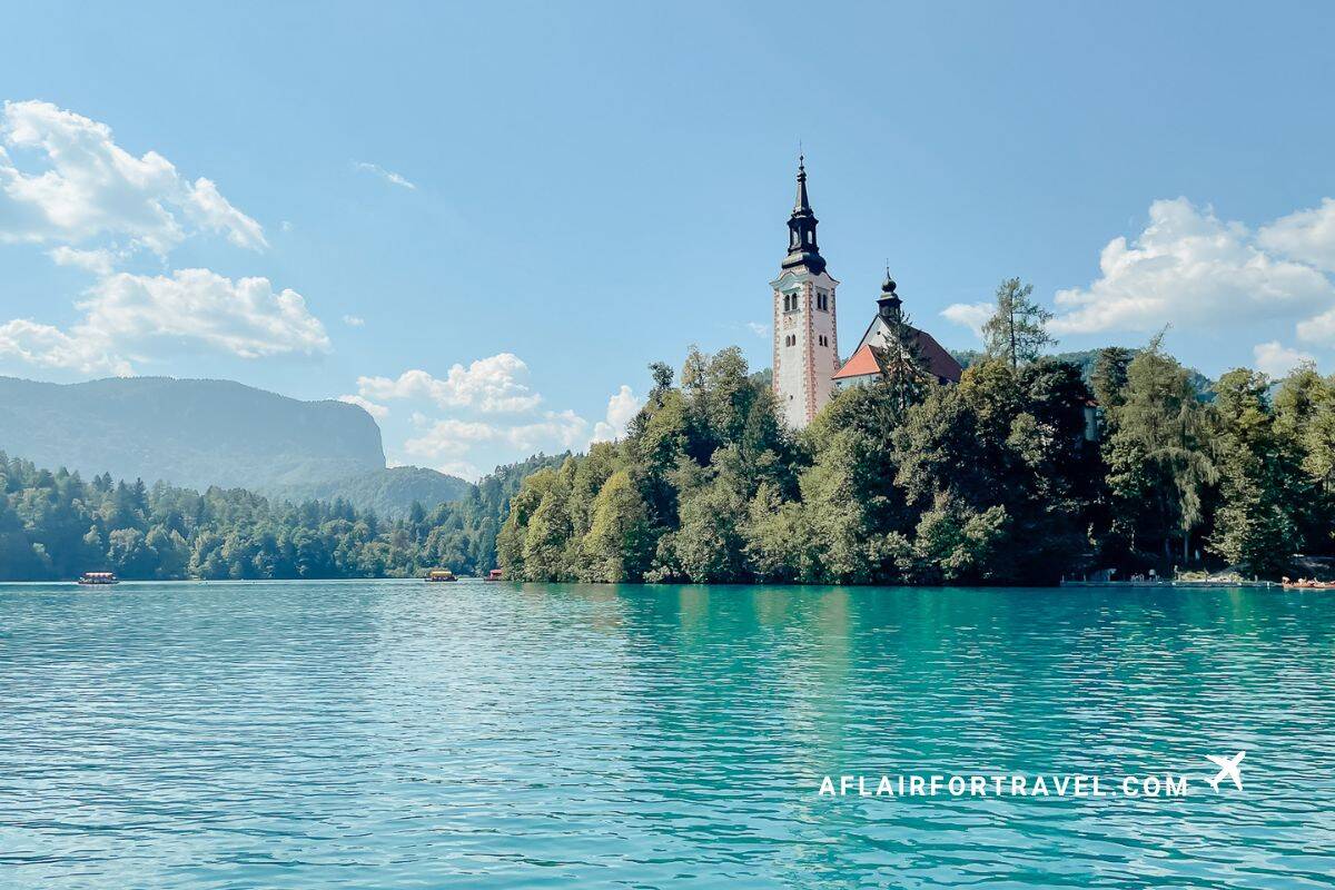 Lake Bled's iconic island with the Church of the Assumption rising from turquoise waters, surrounded by dense forest and Julian Alps mountains under a blue sky with white clouds, one of Croatia's neighboring countries which is worth visiting