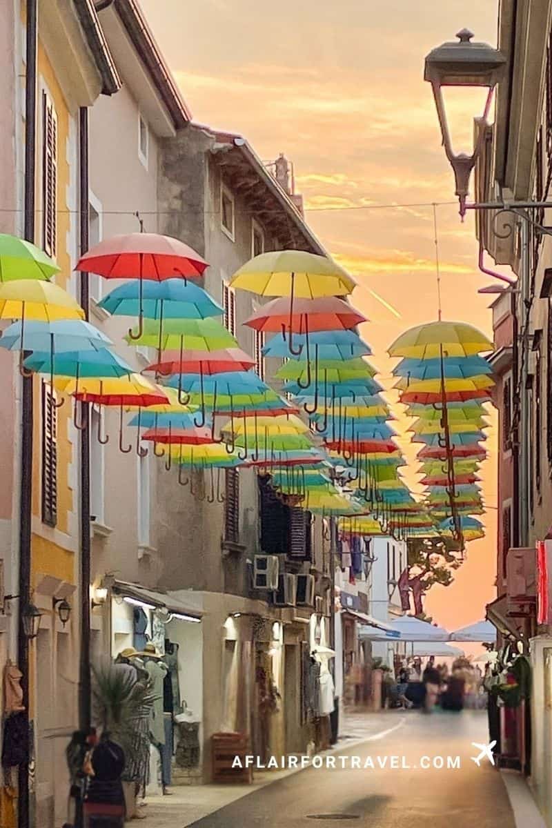 Narrow cobblestone street in Novigrad, Croatia, decorated with hundreds of colorful rainbow umbrellas suspended overhead at sunset, with street lamps and pedestrians below