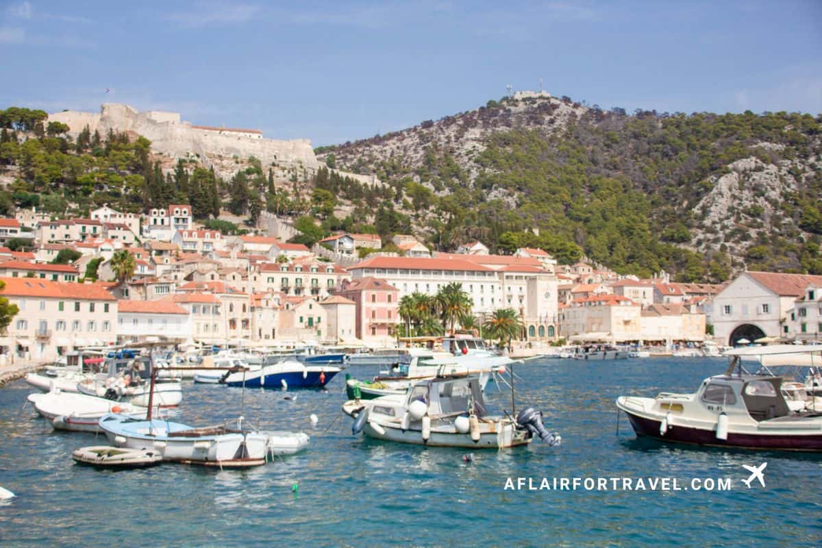 Hvar town harbor filled with fishing boats and yachts, backed by pastel-colored Mediterranean buildings, historic stone fortress on the hillside, and dramatic rocky mountains