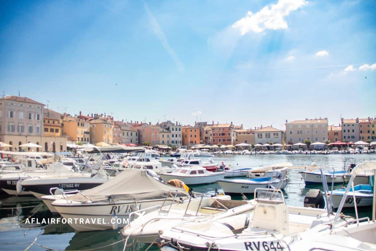 Rovinj harbor packed with moored boats and yachts in the foreground, with colorful pastel buildings of the old town waterfront and beach umbrellas visible across the bay