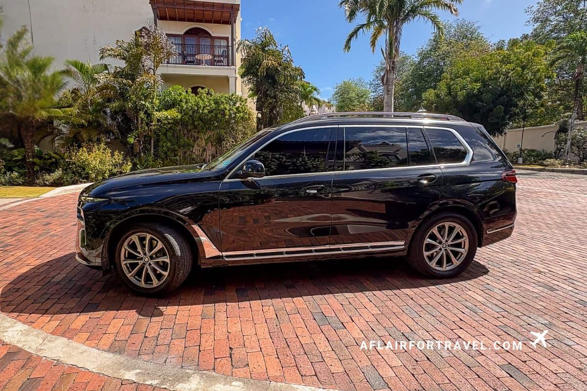 Black luxury BMW X7 SUV parked at Cap Maison resort entrance with palm trees and colonial-style villa in background; used for luxury transfers which is great reason to stay at Cap Maison St. Lucia