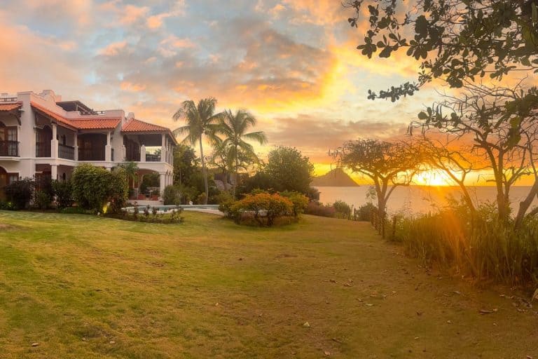 Sunset view at Cap Maison luxury resort in St. Lucia with white colonial-style villa, terracotta roof, manicured lawn, palm trees, and golden sky over Caribbean Sea